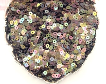 May include: A heart-shaped decorative item covered in iridescent sequins. The sequins are primarily dark brown with hints of green, purple, and gold. The item has a silver-colored scalloped edge and is partially obscured by a pink flower.