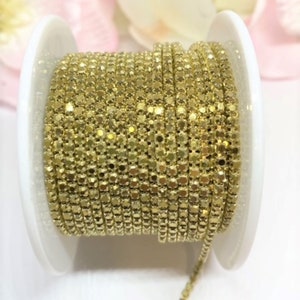 May include: A spool of gold-colored chain, tightly wound and ready for use. The chain is made of small, square-shaped links that catch the light, creating a shimmering effect. The spool has white sides and a central core, providing a clean contrast to the chain.