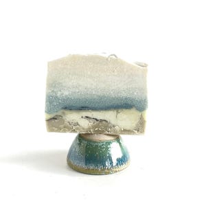 May include: A handmade soap bar with a layered design in white, blue, and beige, displayed on a small, glazed ceramic pedestal. The pedestal is a rounded shape with a green and blue colour scheme.