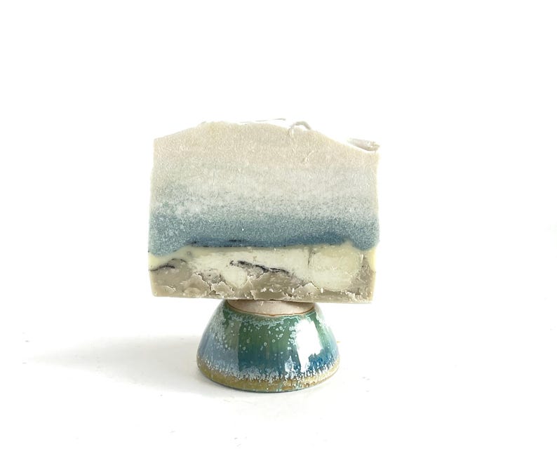 May include: A handmade soap bar with a layered design in white, blue, and beige, displayed on a small, glazed ceramic pedestal. The pedestal is a rounded shape with a green and blue colour scheme.