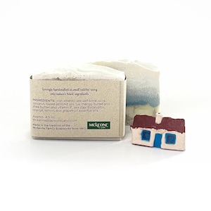 May include: Two bars of handmade soap and a small decorative house. One soap bar is white with a blue top, the other is white with a beige box. The box has text about ingredients and the maker.