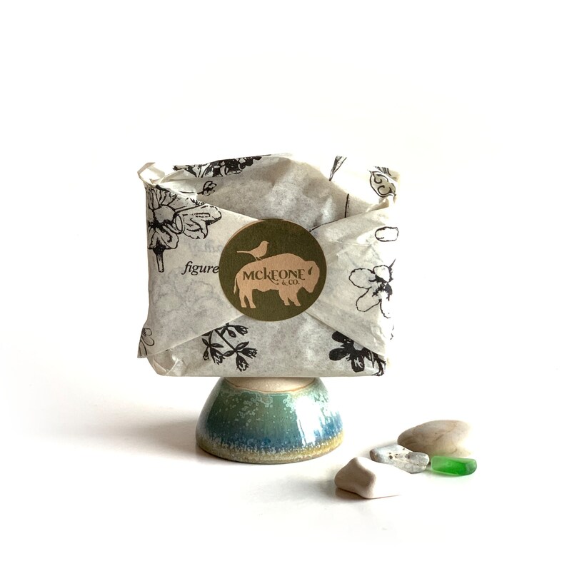May include: A small, wrapped parcel on a ceramic pedestal. The parcel is wrapped in floral-printed paper and sealed with a round sticker featuring a bison and the text "MCKEONE & CO.". Several small stones and a piece of green glass are next to the pedestal.