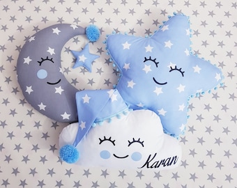 May include: Three plush pillows shaped like a crescent moon, a star, and a cloud. The moon is gray with white stars, the star is blue with white stars, and the cloud is white with a blue sleeping cap. All three pillows have smiling faces with eyelashes and blue eyes. The cloud pillow has the name "Karan" embroidered on it.