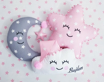 May include: Three plush toys in the shape of a crescent moon, a star, and a cloud. The moon is gray with white stars and a pink cheek. The star is pink with white stars and a smiling face. The cloud is white with a pink hat and a smiling face. The toys are on a white background with pink stars.