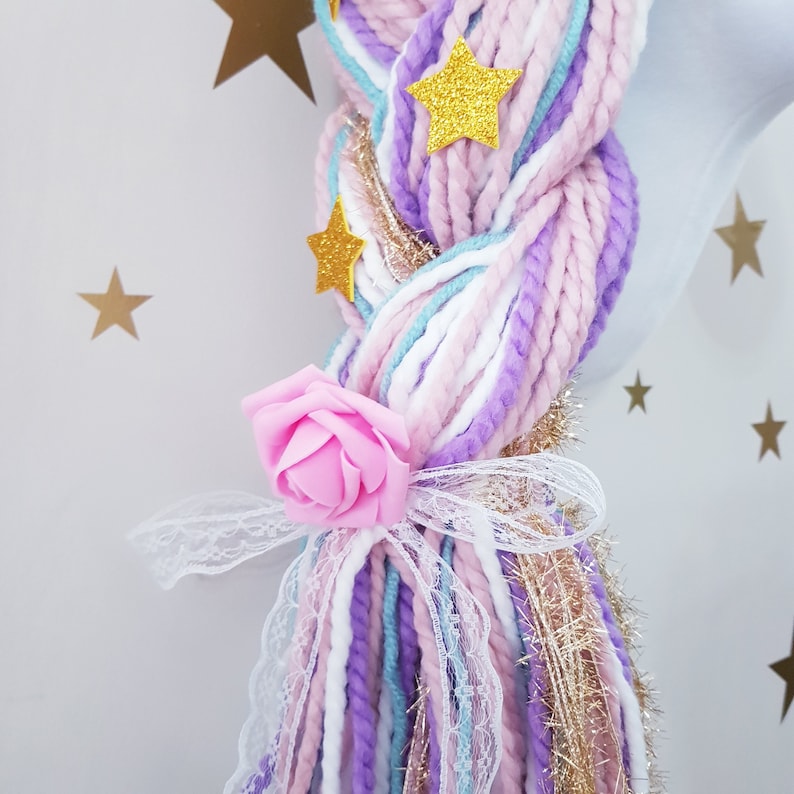 May include: A close-up of a unicorn mane with a pink rose and gold star accents. The mane is braided with strands of white, light blue, lavender, and pink yarn. A white lace bow and gold tinsel add detail.