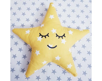 May include: A yellow star-shaped pillow with a white star pattern and a smiling face with closed eyes. The pillow is on a white background with a gray star pattern.