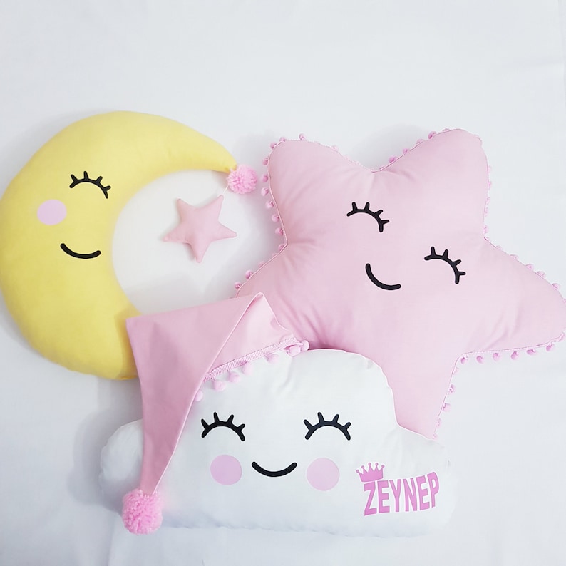 May include: Three plush pillows shaped like a crescent moon, a star, and a cloud. The moon is yellow, the star is pink, and the cloud is white. Each pillow has a smiling face with eyelashes and pink cheeks. The cloud pillow has a pink sleeping cap and the text "ZEYNEP" on the bottom.