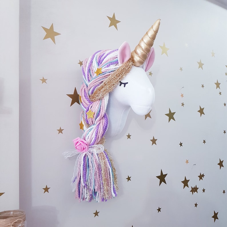May include: A white unicorn head wall decoration with a gold horn and pink ears. The unicorn has a long, braided mane made of purple, pink, and blue yarn, decorated with gold stars and a pink rose. The wall is decorated with gold star stickers.