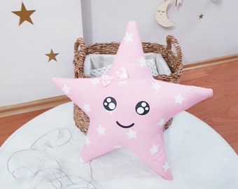 May include: Pink star-shaped pillow with a white star pattern and a smiling face. The pillow has a pink bow and black eyes.