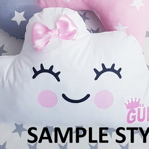 May include: White cloud-shaped pillow with a pink bow, black eyelashes, a smiling mouth, and pink cheeks. The pillow has the text "GULCE" in pink letters with a crown above it. The pillow is on a gray background with white stars.