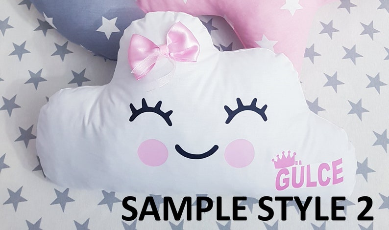 May include: White cloud-shaped pillow with a pink bow, black eyelashes, a smiling mouth, and pink cheeks. The pillow has the text "GULCE" in pink letters with a crown above it. The pillow is on a gray background with white stars.