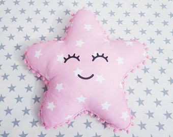 May include: A pink star-shaped pillow with a smiling face and white stars on it. The pillow has a pom-pom trim around the edges.