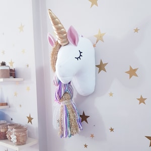 May include: A white unicorn head wall decoration with a gold horn, pink ears, and a multi-coloured braid. The unicorn has closed eyes with black eyelashes. The wall is decorated with gold star stickers.
