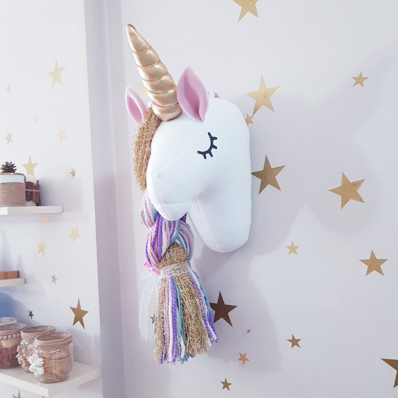 May include: A white unicorn head wall decoration with a gold horn, pink ears, and a multi-coloured braid. The unicorn has closed eyes with black eyelashes. The wall is decorated with gold star stickers.