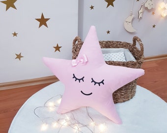 May include: A pink star-shaped pillow with a smiling face and closed eyes. The pillow has a small pink bow on top and is sitting on a white fluffy rug with string lights.