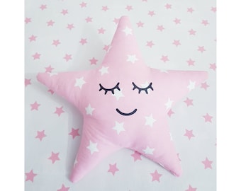 May include: A pink star-shaped pillow with a white star pattern and a smiling face with closed eyes. The pillow is on a white background with pink stars.