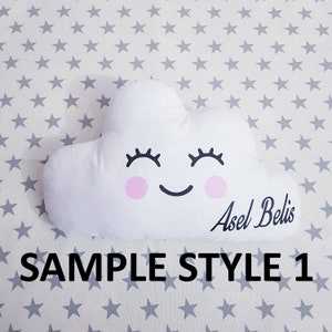 May include: White cloud-shaped pillow with a smiling face, pink cheeks, and eyelashes. The pillow has the name "Asel Belis" written on it. The pillow is on a white background with gray stars.
