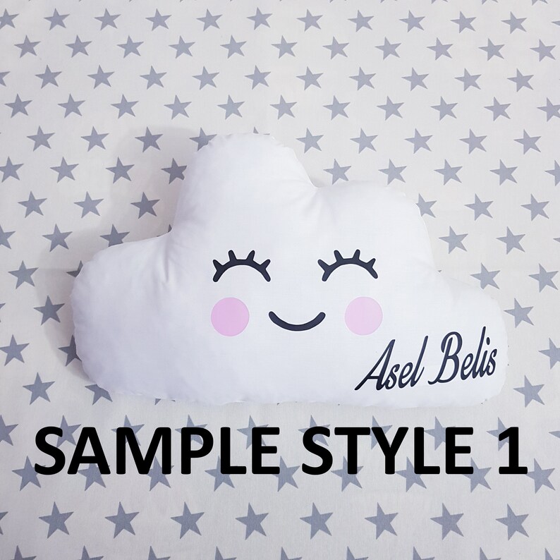 May include: White cloud-shaped pillow with a smiling face, pink cheeks, and eyelashes. The pillow has the name "Asel Belis" written on it. The pillow is on a white background with gray stars.