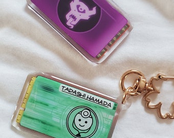 May include: Two keychains, one purple with a black and white demon design and the text "Tadashi Hamada" and the other green with a white cartoon robot with a stethoscope.