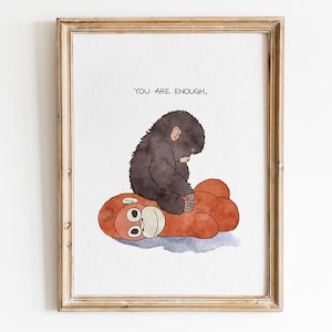 May include: Watercolor print in a wooden frame. A small, dark-furred gorilla sits atop a red and orange toy. The text "YOU ARE ENOUGH" is above the gorilla. The artwork has a whimsical and comforting theme.
