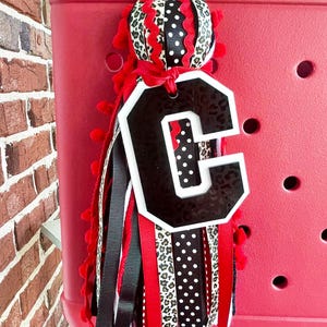 May include: A red, white and black letter C charm with a leopard print, polka dot and red ribbon tassel. The charm is attached to a red tote bag.