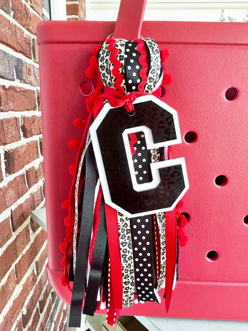 May include: A red, white and black letter C charm with a leopard print, polka dot and red ribbon tassel. The charm is attached to a red tote bag.