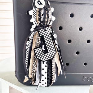 May include: A black and white decorative bag charm with a checkered letter "J" and a white pom-pom accent. The charm is attached to a black bag. The charm features ribbons with polka dots, leopard print, and lace.