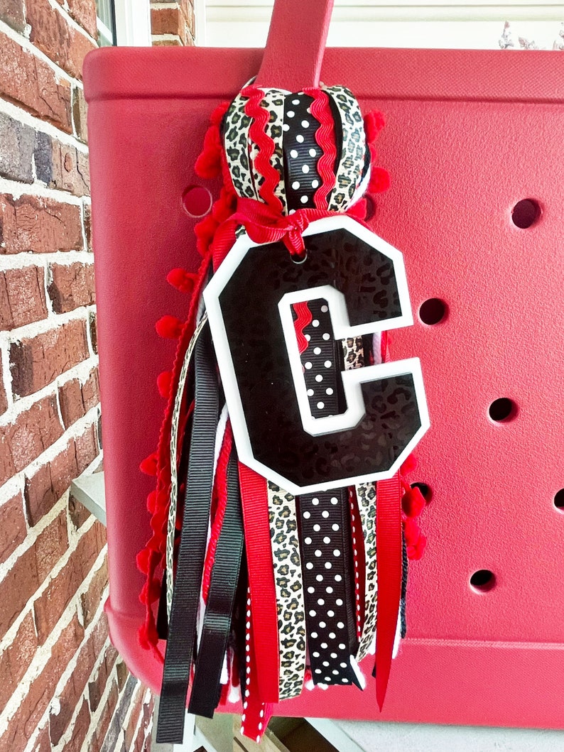 May include: A red, white and black monogrammed charm with a letter "C" in a white outline. The charm is attached to a red, black and white ribbon with a leopard print and polka dot pattern. The charm is hanging from a red tote bag.
