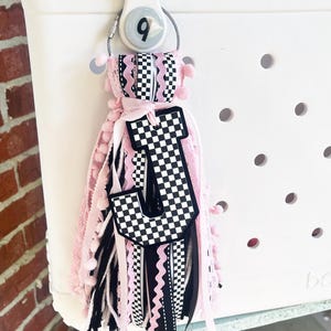 May include: A pink and black checkered letter J charm with a black and white ribbon tassel attached to a white tote bag.