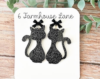 May include: Black glitter cat earrings with black bows, displayed on a white card. The card reads "6 Farmhouse Lane" and "6farmhouselane.etsy.com or on Facebook". The earrings have a cat silhouette design.