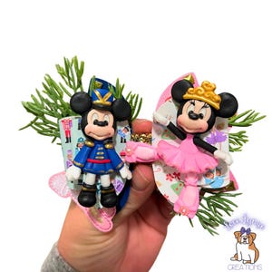 May include: A pair of handmade hair bows featuring a nutcracker-themed design. One bow features a pink ballerina Minnie Mouse with a gold crown and the other features a blue and red Mickey Mouse dressed as a nutcracker soldier.