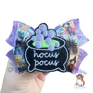 May include: A large, decorative hair bow featuring a black cauldron with the words "hocus pocus" embroidered in white. The bow has a purple glittery base and a patterned center with images of witches. A small logo is visible.