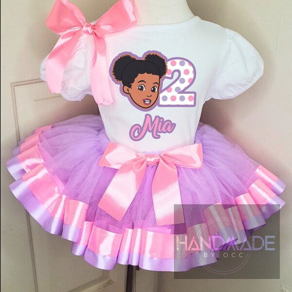 May include: A white t-shirt with a pink bow, a cartoon girl with brown hair, and the number 2 with pink and purple polka dots. The shirt says "Mia" in purple letters. The shirt is paired with a purple tulle tutu with pink satin ribbon trim.
