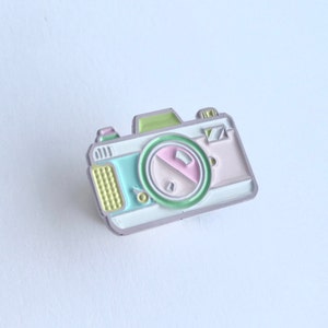 May include: A pastel-colored enamel pin shaped like a vintage camera. The camera is light pink, light blue, light green, and yellow. The lens is a light green circle with a pink and white circle inside.