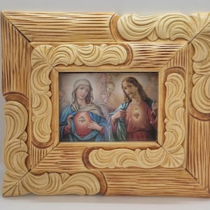 May include: A rectangular wooden frame with a religious image. The frame has a light brown color and ornate, carved details. The image depicts two figures with halos, one in blue and the other in red, with a heart symbol.