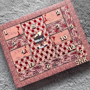 Peut inclure: Un calendrier de l'avent rectangulaire dans des tons rose et rouge, doté de douze tiroirs numérotés. Le design présente des motifs de feuilles, des pois et une image centrale avec le texte japonais 進撃の巨人. Les lettres SNK sont fixées en bas à droite.