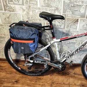 Puede incluir: Una bicicleta de montaña blanca y roja con neumáticos negros y una bolsa de pannier negra y azul unida al portabultos trasero. La bicicleta tiene un manillar negro con una campana roja y un sillín negro y rojo.