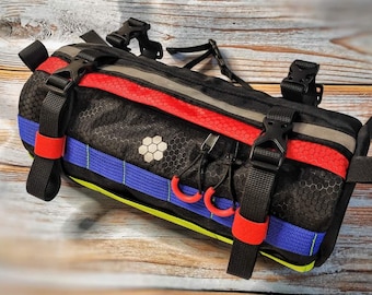 May include: Black bike handlebar bag with red, blue, and yellow accents. The bag has multiple straps and buckles for attaching to a bike.