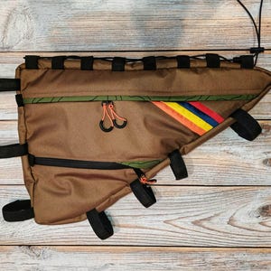 Puede incluir: Bolsa de cuadro de bicicleta marrón con correas y cremalleras negras. La bolsa tiene un diseño de rayas coloridas con detalles en naranja, amarillo y azul. Diseñada para encajar dentro del cuadro de una bicicleta.