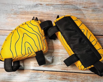 Op de afbeelding: Twee gele fietstasjes voor het stuur met zwarte banden en een zwart gaaspaneel. Eén tasje heeft een golvend patroon en de andere is effen geel.