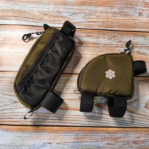 May include: Two green bike bags with black straps and a honeycomb logo. The bag on the left has a mesh pocket and a zipper closure. The bag on the right has a single strap and a buckle closure.