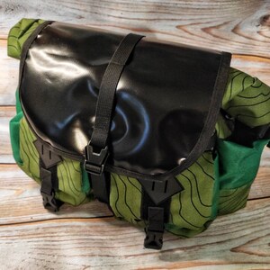 May include: A green and black messenger bag with a waterproof flap. The bag has a topographic pattern and black straps.