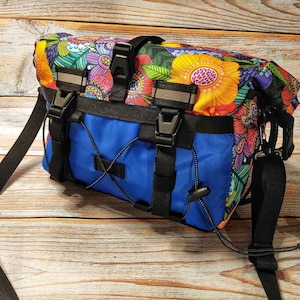Könnte beinhalten: Eine blaue Fahrradtasche mit floralem Muster, schwarzen Riemen und Schnallen. Die Tasche hat einen Reißverschluss und einen Kordelzug.