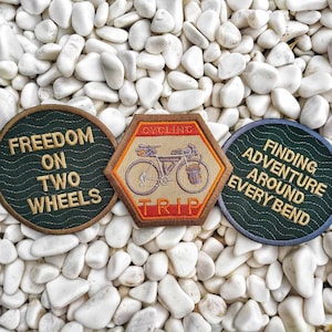Op de afbeelding: Drie geborduurde patches met fietsmotieven. De eerste patch is groen met witte tekst die "Freedom on Two Wheels" luidt. De tweede patch is oranje, bruin en wit met een fiets-illustratie en de tekst "Cycling Trip". De derde patch is groen met witte tekst die "Finding Adventure Around Every Bend" luidt.