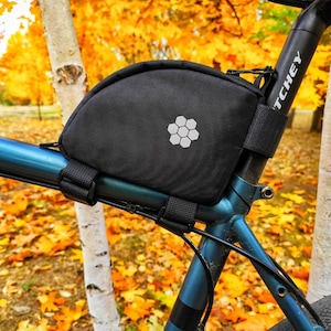 Puede incluir: Una bolsa de asiento de bicicleta negra con un logotipo de panal de abeja unida a un cuadro de bicicleta azul. La bicicleta está aparcada junto a un abedul con un fondo de follaje otoñal.