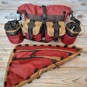 Może przedstawiać: Zestaw czerwonych i beżowych toreb i akcesoriów rowerowych. Zestaw zawiera dużą torbę na kierownicę, dwa uchwyty na butelki z wodą i torbę na ramę. Torby mają czarne paski i klamry.