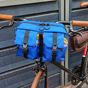 Op de afbeelding: Zwarte fiets met bruine banden, stuur en zadel. Een blauwe stuurtas met zwarte riemen en gespen is aan de voorkant bevestigd. Een bruine tas is onder het zadel gemonteerd. De fiets staat op een bakstenen oppervlak.