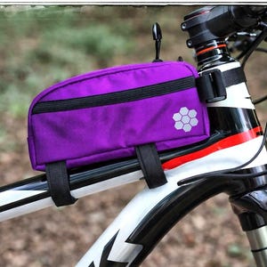 以下が含まれることがあります： 紫色のハンドルバーバッグが付いた自転車のフレームのクローズアップ。バッグには黒いジッパーと六角形のデザインがあります。自転車のフレームは白、黒、赤です。