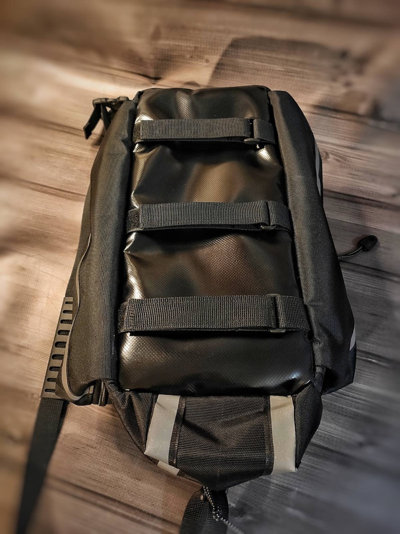 May include: Black bicycle pannier bag with reflective stripes and adjustable straps. The bag is made of a durable, waterproof material and features multiple compartments for storage. The bag is designed to attach to a bicycle rack for easy transport of items.