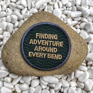 Peut inclure: Un patch brod&eacute; vert et gris avec le texte "FINDING ADVENTURE AROUND EVERY BEND" en fil dor&eacute;. Le patch est sur un rocher blanc.